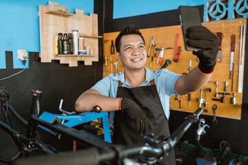a handsome repairman smiling in an apron while taking a selfie using a cellphone while repairing a bicycle in a bicycle repair shop