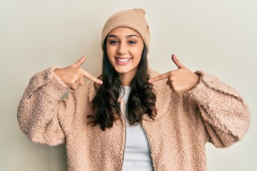 Young hispanic woman wearing wool sweater and winter hat looking confident with smile on face, pointing oneself with fingers proud and happy.