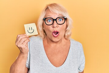 Middle age blonde woman holding smile reminder scared and amazed with open mouth for surprise, disbelief face
