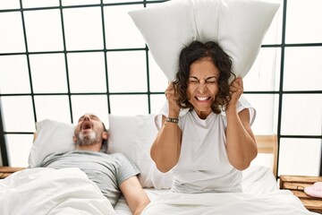 Middle age hispanic man snoring and woman angry covering ears with pillow at bedroom.
