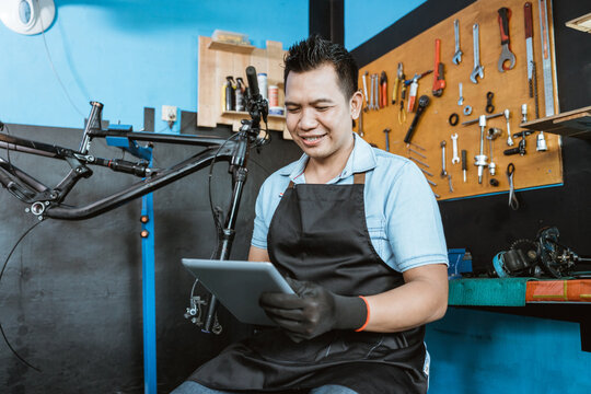 a handsome bicycle mechanic uses a digital tablet to browse against the background of a bicycle frame in a workshop
