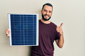 Young man with beard holding photovoltaic solar panel smiling happy and positive, thumb up doing...