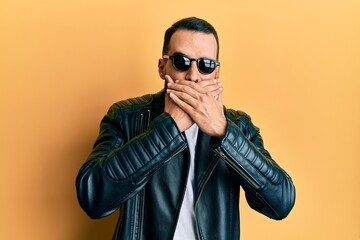 Obraz premium Young man with beard wearing black leather jacket and sunglasses shocked covering mouth with hands for mistake. secret concept.