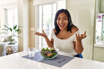 Obraz na płótnie Canvas Young hispanic woman eating healthy salad at home celebrating crazy and amazed for success with arms raised and open eyes screaming excited. winner concept