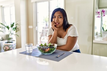 Obraz na płótnie Canvas Young hispanic woman eating healthy salad at home looking stressed and nervous with hands on mouth biting nails. anxiety problem.