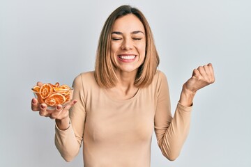 Young caucasian blonde woman holding bowl of dry orange screaming proud, celebrating victory and success very excited with raised arm