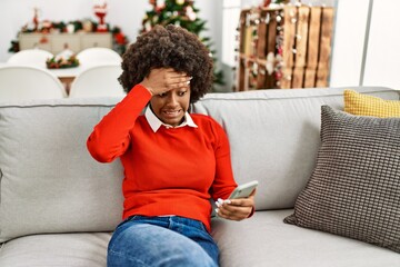 Young african american woman with afro hair using smartphone sitting on the sofa by christmas tree...