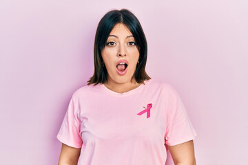 Fototapeta premium Young hispanic woman wearing pink cancer ribbon on t shirt in shock face, looking skeptical and sarcastic, surprised with open mouth