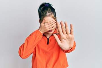 Young modern girl wearing sports sweatshirt covering eyes with hands and doing stop gesture with...