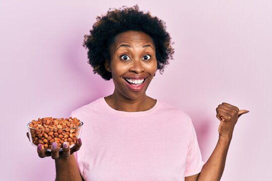 Young african american woman holding peanuts pointing thumb up to the side smiling happy with open mouth