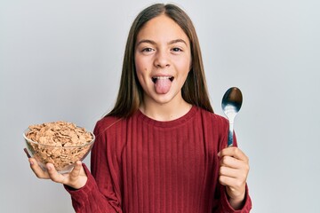 Beautiful brunette little girl eating healthy whole grain cereals holding spoon sticking tongue out...