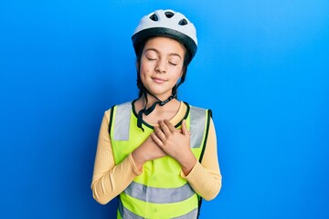 Beautiful brunette little girl wearing bike helmet and reflective vest smiling with hands on chest with closed eyes and grateful gesture on face. health concept.