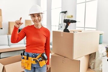 Middle age grey-haired woman wearing hardhat standing at new home smiling and confident gesturing with hand doing small size sign with fingers looking and the camera. measure concept.