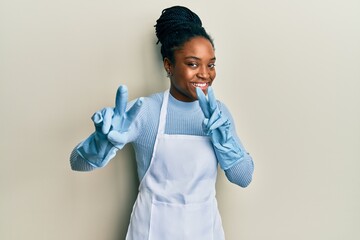 African american woman with braided hair wearing cleaner apron and gloves smiling looking to the...