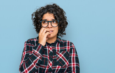 Fototapeta na wymiar Young hispanic woman with curly hair wearing casual clothes and glasses looking stressed and nervous with hands on mouth biting nails. anxiety problem.