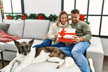 Young caucasian couple smiling happy unboxing gift sitting on the sofa with dog by christmas tree at home.