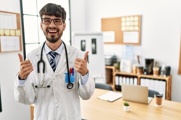 Hispanic man with beard wearing doctor uniform and stethoscope at the office success sign doing positive gesture with hand, thumbs up smiling and happy. cheerful expression and winner gesture.