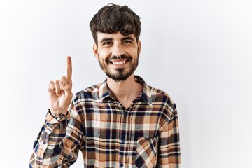 Fototapeta premium Hispanic man with beard standing over isolated background showing and pointing up with finger number one while smiling confident and happy.