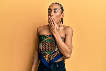 Hispanic man wearing make up and long hair wearing womans top bored yawning tired covering mouth with hand. restless and sleepiness.