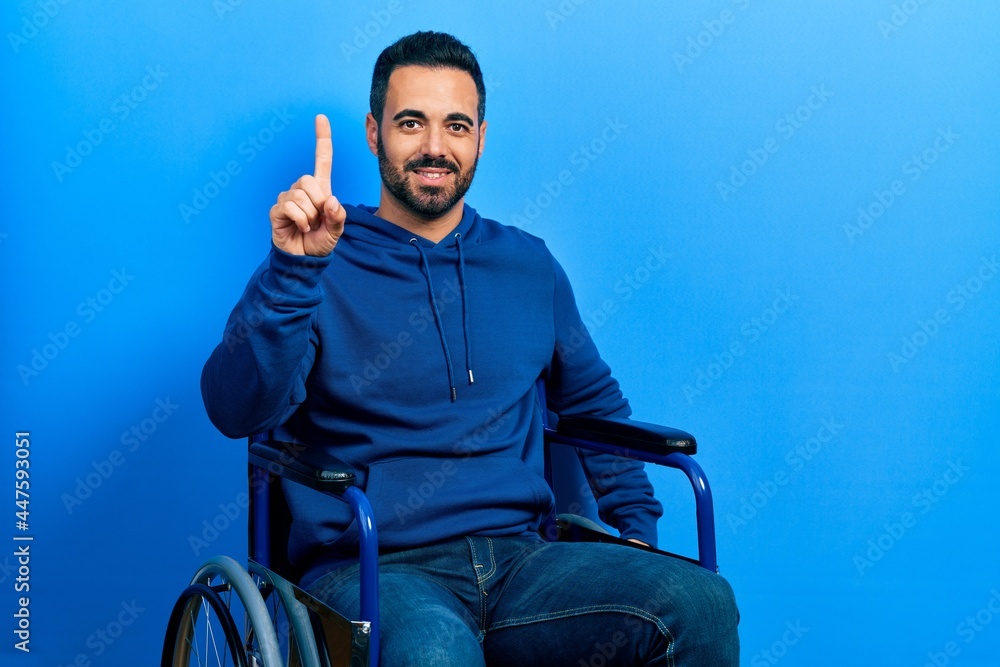 Sticker handsome hispanic man with beard sitting on wheelchair showing and pointing up with finger number on