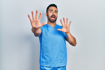 Handsome hispanic man with beard wearing blue male nurse uniform afraid and terrified with fear...