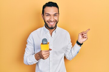 Hispanic man with beard holding reporter microphone smiling happy pointing with hand and finger to...