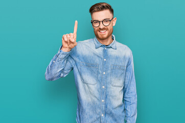 Young redhead man wearing casual denim shirt showing and pointing up with finger number one while smiling confident and happy.