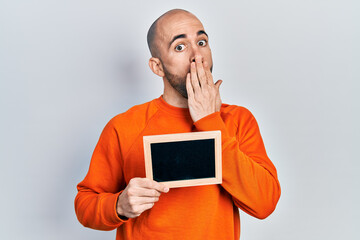 Young bald man holding blackboard covering mouth with hand, shocked and afraid for mistake....
