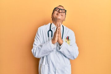 Middle age indian man wearing doctor coat and stethoscope begging and praying with hands together...