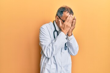 Middle age indian man wearing doctor coat and stethoscope with sad expression covering face with...