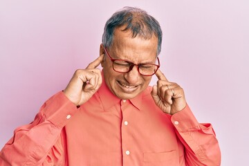 Middle age indian man wearing casual clothes and glasses covering ears with fingers with annoyed expression for the noise of loud music. deaf concept.