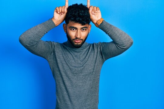 Arab man with beard wearing turtleneck sweater doing funny gesture with finger over head as bull horns