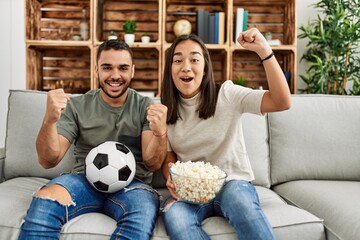 Young latin couple watching soccer match eating porpcorn at home.