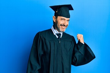 Middle age hispanic man wearing graduation cap and ceremony robe smiling with happy face looking and pointing to the side with thumb up.