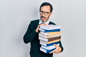 Middle age hispanic man holding a pile of books in shock face, looking skeptical and sarcastic,...