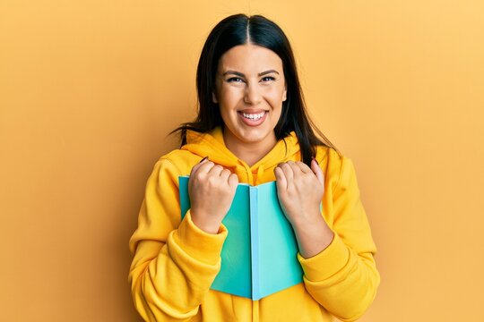 Beautiful brunette woman holding book smiling with a happy and cool smile on face. showing teeth.