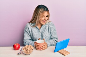 Young caucasian woman having breakfast looking at the tablet sitting on the tablet smiling and laughing hard out loud because funny crazy joke.
