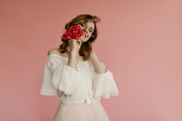 Stylish girl with curly hair in white trendy blouse smiling on isolated background. Woman in good mood holding pink flowers..