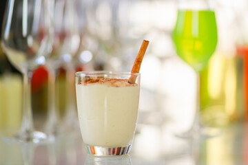 Close up shot of creamy spiced eggnog with gingerbread in glass on decorate Thanksgiving Christmas eve party table full of empty tall wine glasses and other alcoholic beverages in blurred background
