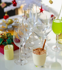Tasty delicious milk cream eggnog drink with gingerbread in glass on table in front empty tall wine glasses present gift boxes white chocolate candles green apple punch and decorated Christmas tree