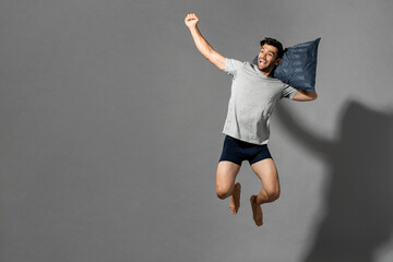 Fresh and energetic young Caucasian man in sleepwear jumping with pillow after wake up in light...