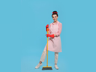 Young housewife with broom on light blue background, space for text