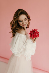 Joyful woman with wavy hairstyle in white fluffy dress smiling and holding bright bouquet on pink background. Cool girl in light outfit posing..
