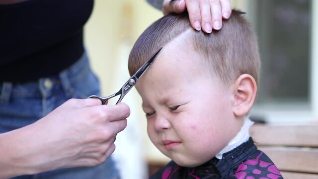 portrait of a little baby boy with closed eyes and scaring emotions while hairdresser cuts his fringe or bangs with scissors. close-up front view