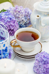 A visual for content. Still life in vintage style. A mug with a drink, an old book, a cage, candles and hydrangea flowers in the garden on a white wooden table. The concept of a tea ceremony.