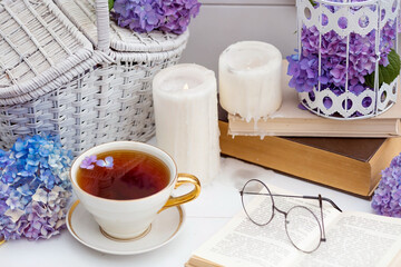 A visual for content. Still life in vintage style. A mug with a drink, an old book, a cage, candles and hydrangea flowers in the garden on a white wooden table. The concept of a tea ceremony.
