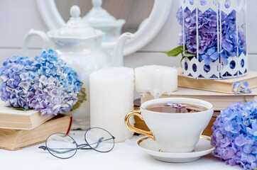 A visual for content. Still life in vintage style. A mug with a drink, an old book, a cage, candles and hydrangea flowers in the garden on a white wooden table. The concept of a tea ceremony.