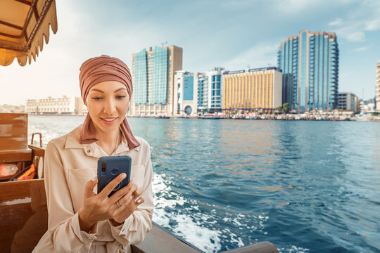 Happy asian woman in a maroon turban using app on her smartphone while cruising on a traditional Abra Dhow boat on Dubai Creek.
