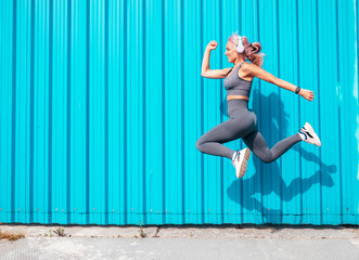 Fitness smiling woman in grey sports clothing with pink hair. Young beautiful model with perfect body.Female jumping and jogging in street near blue wall.Listening music in wireless headphones