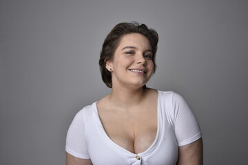 Close up portrait of young plus sized woman with short brunette hair,  wearing a white shirt, with over the top emotional facial expressions against a light studio background.  